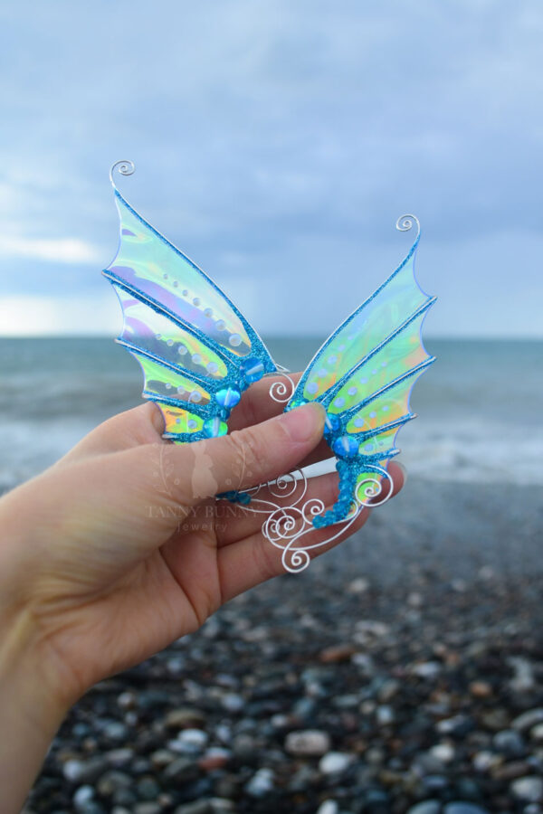 Hand holding a pair of handmade blue and gold mermaid ear cuffs with white glitter in front of the sea