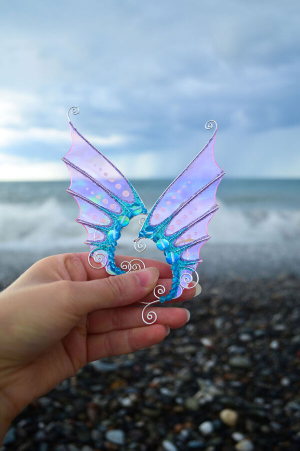 Hand holding pink and blue mermaid ear cuffs with sea background