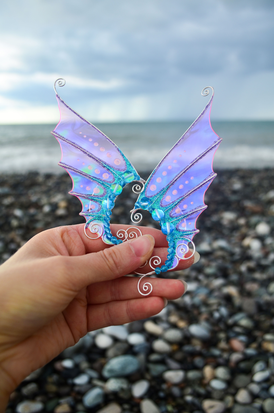 Pink and blue mermaid ear cuffs held in hand against a seashore background, handmade cosplay accessory