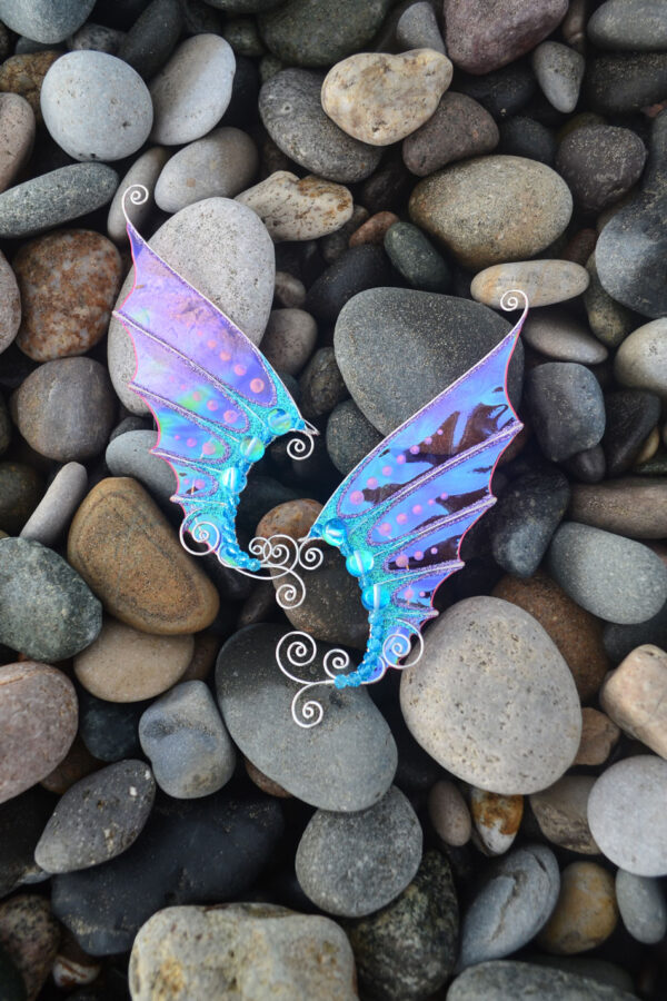 Pair of handmade pink and blue mermaid ear cuffs with glitter placed on smooth beach stones