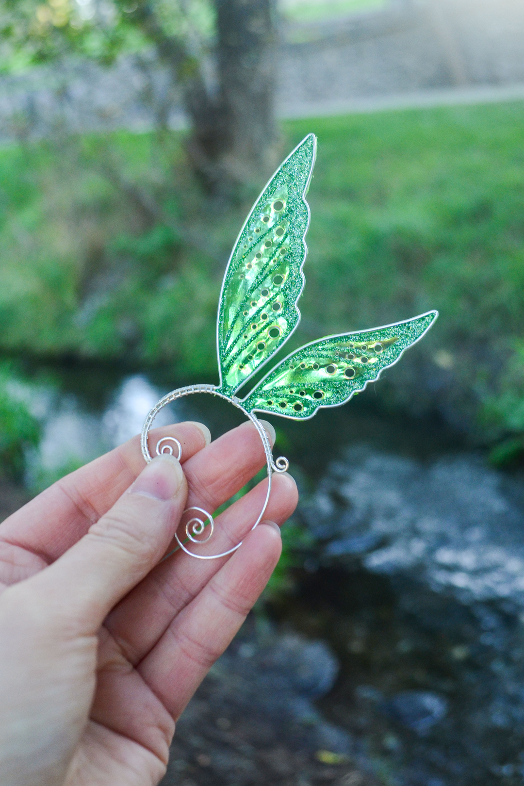 Fairy cosplay ear cuff, green, handmade, no piercing needed, fairy earring — green iridescent wing-shaped ear cuff held in a hand outdoors near a stream.