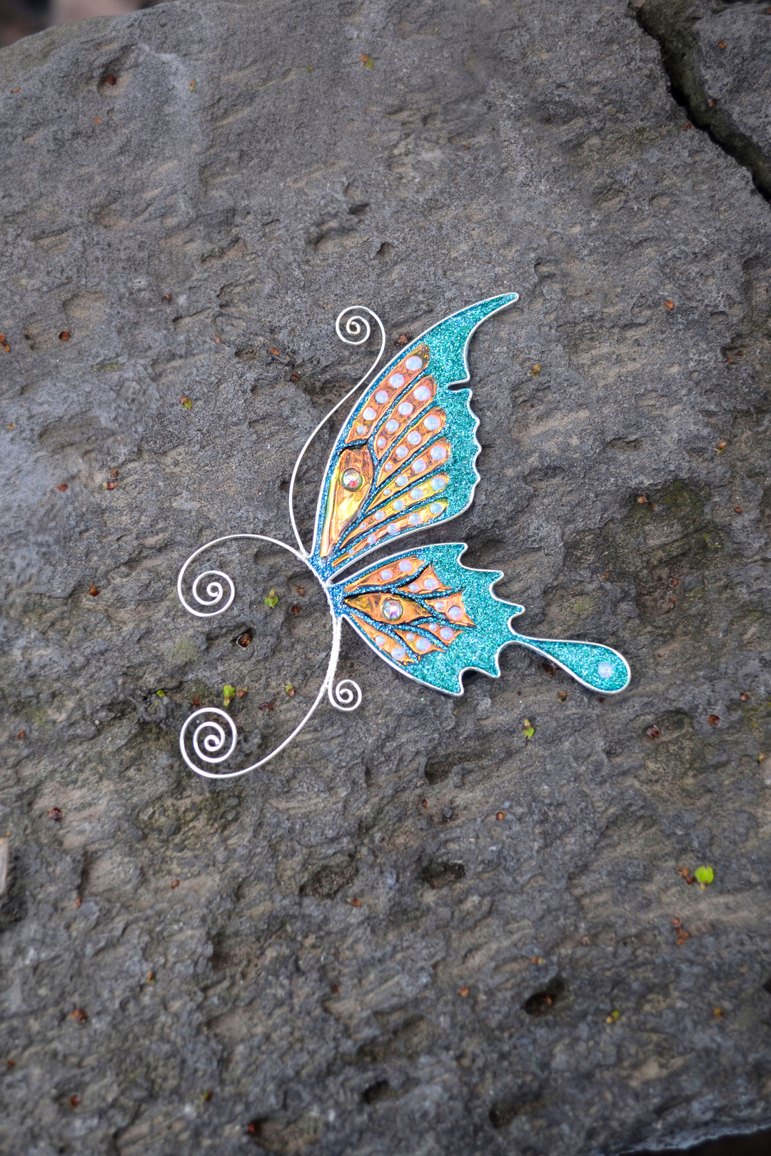 Handmade butterfly ear cuff placed on grey stone surface, teal and gold fairy wing earring