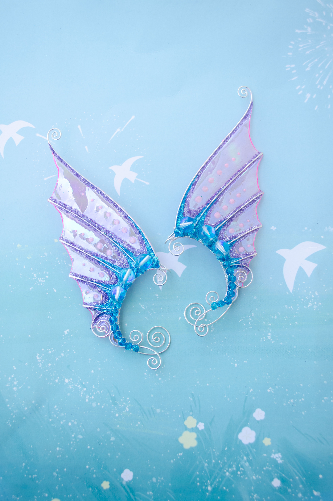 Pink and blue mermaid ear cuffs on pastel blue background with decorative elements, handmade no-piercing jewelry