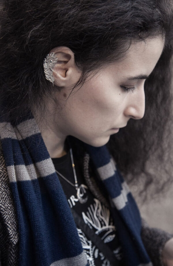 Silver feather ear cuff with blue stones worn on the right ear, shown on a woman with dark curly hair and a navy scarf.