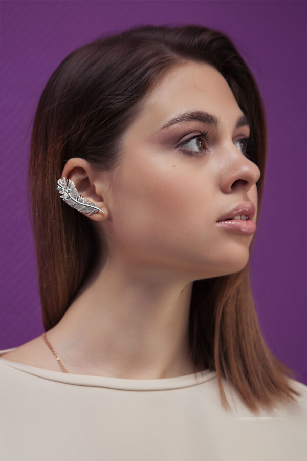 Silver feather ear cuff with blue stones worn on right ear, shown on model with brown hair and cream-colored blouse against purple background.