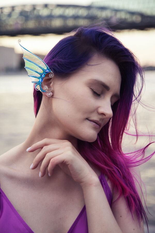 Model with vibrant hair wearing a handmade blue and gold mermaid ear cuff with white glitter near water at sunset