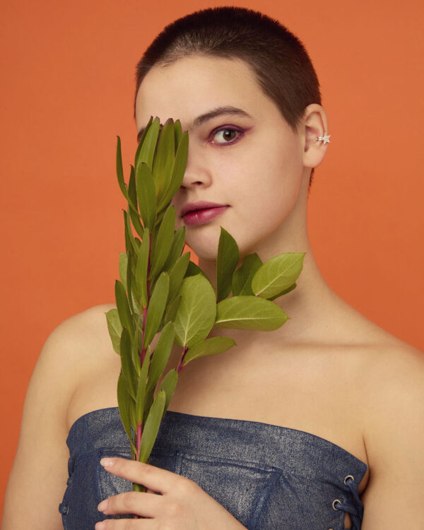 Model with shaved head and purple eyeliner wearing a silver star ear cuff, holding green leaves in front of face.
