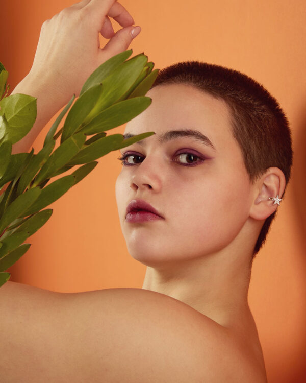 Model with buzz cut and purple eyeshadow wearing a silver star ear cuff with clear stones, holding green leaves.