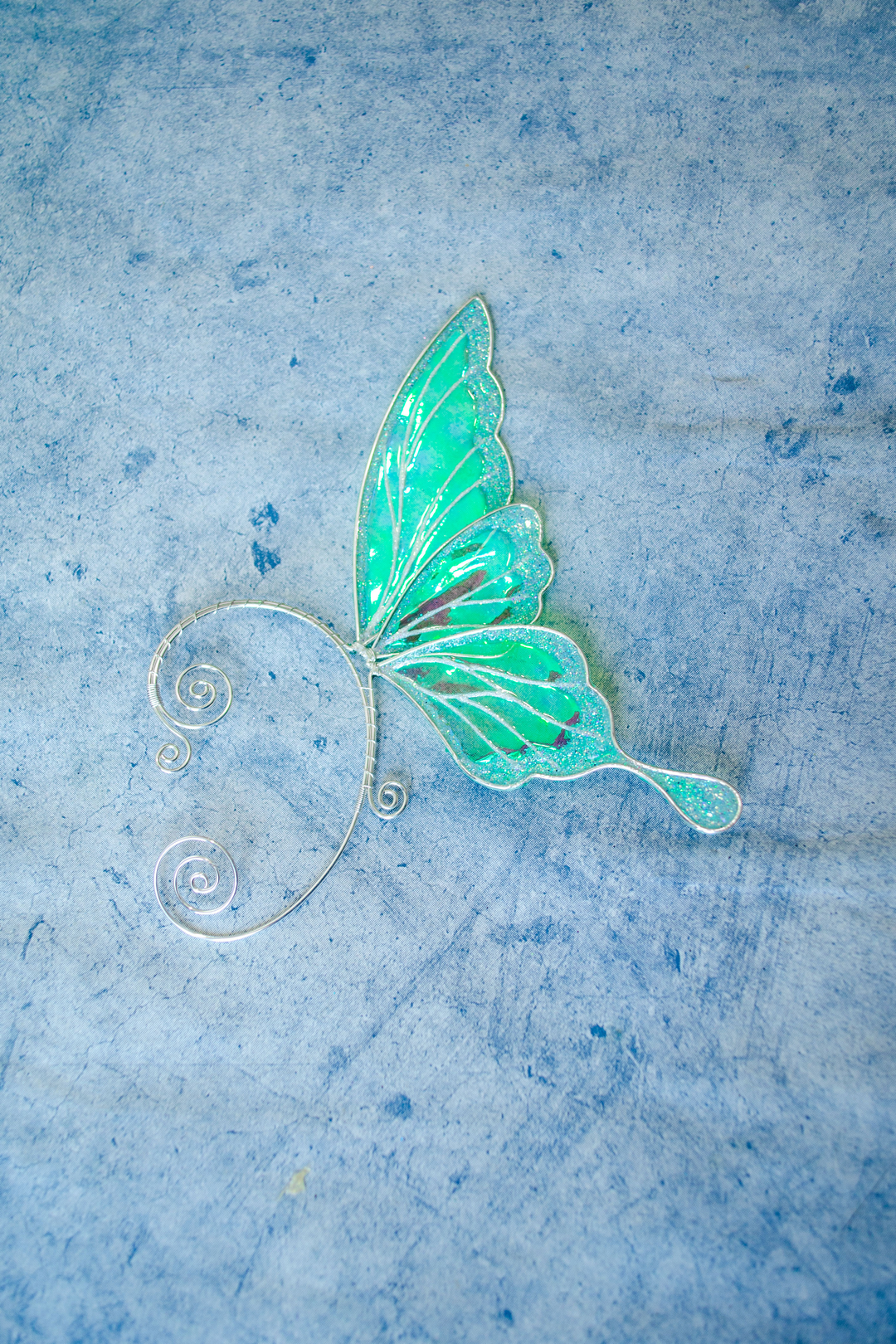 iridescent fairy wing ear cuff in green and white on grey textured background, handmade cosplay accessory